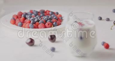 将焦点从一个带有成熟甜树莓蓝莓的盘子中转移，樱桃放在一个带有浆果和牛奶的杯子上，放在白色的桌子上